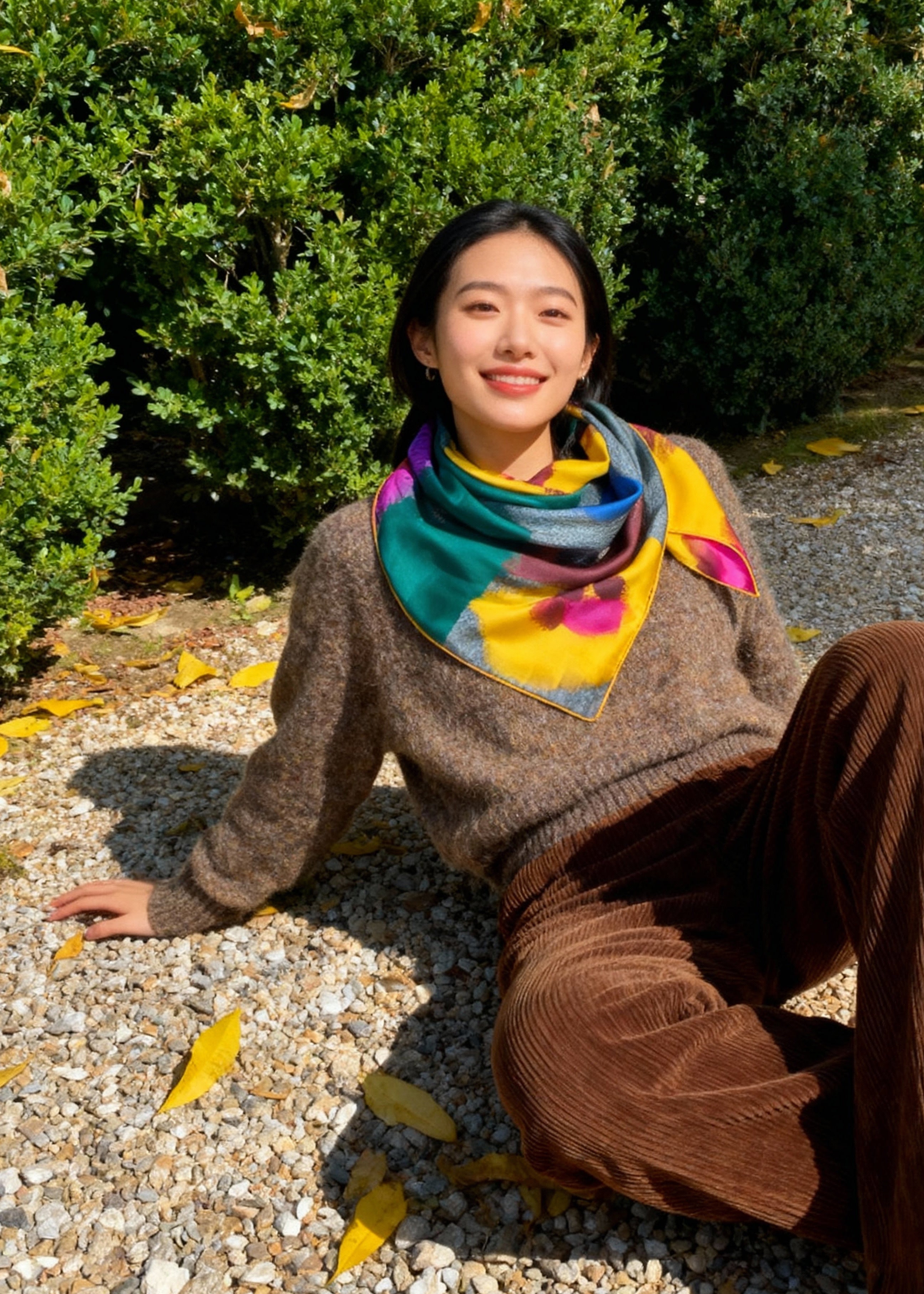 Person sitting on a gravel path with green bushes, she wears a floral print silk square around the neck