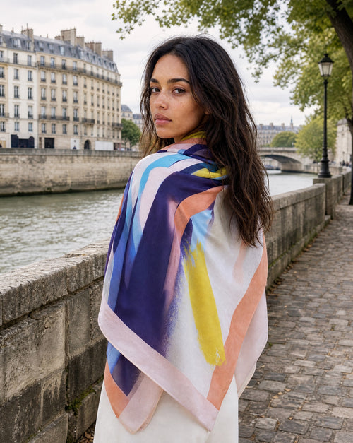 model wears square over her shoulders, the print is geometric abstract with urple , yellow, roa shades